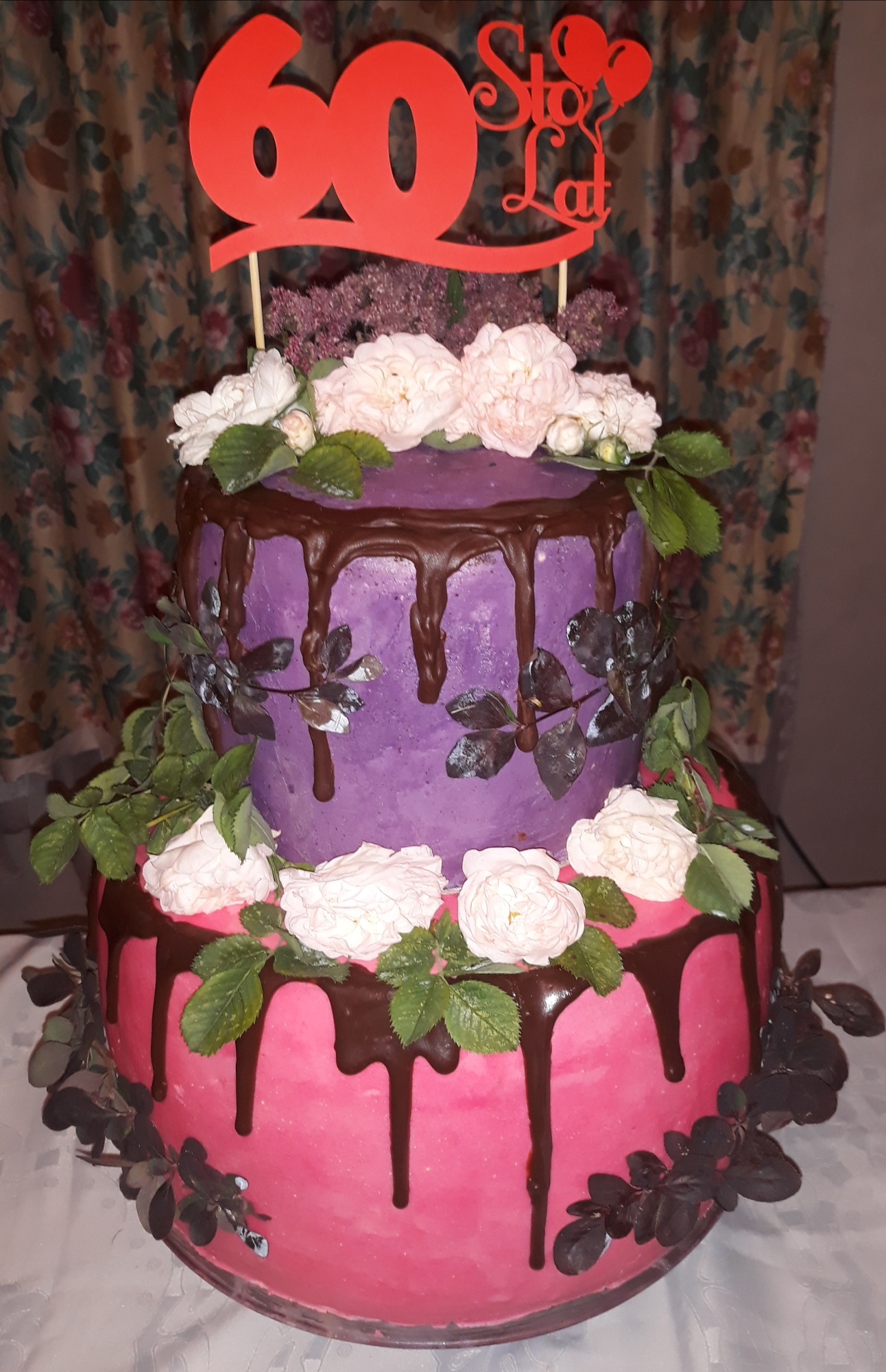 Dwu-piętrowy tort urodzinowy z różowym i fioletowym kremem, dekorowany czekoladowymi polewami, białymi różami i topperem z napisem '60 Sto Lat'.