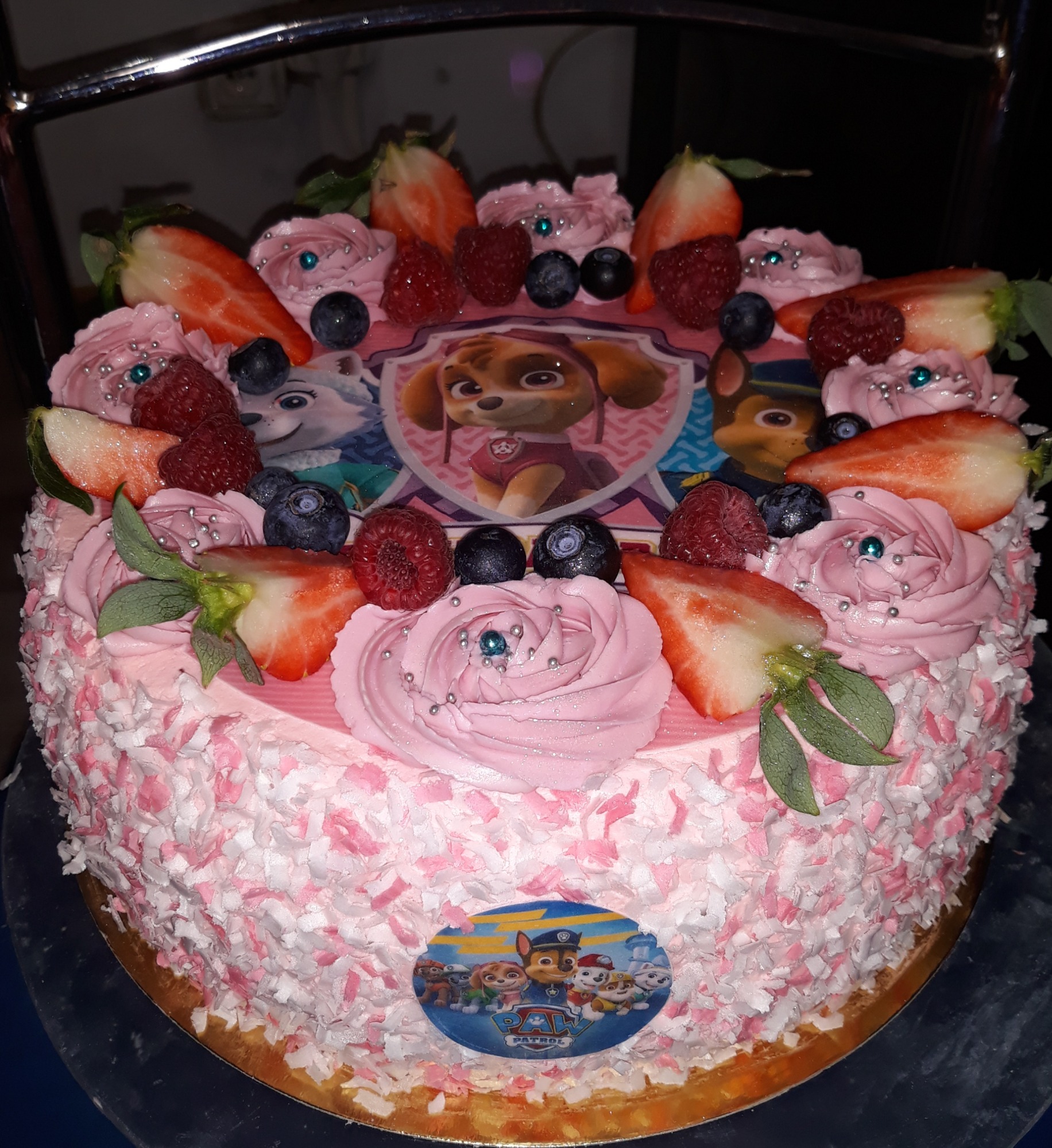 Okrągły tort urodzinowy z różowym kremem, posypką z białych i różowych wiórków, dekorowany truskawkami, malinami, borówkami, kremowymi rozetkami z srebrnymi koralikami i jadalnymi obrazkami...