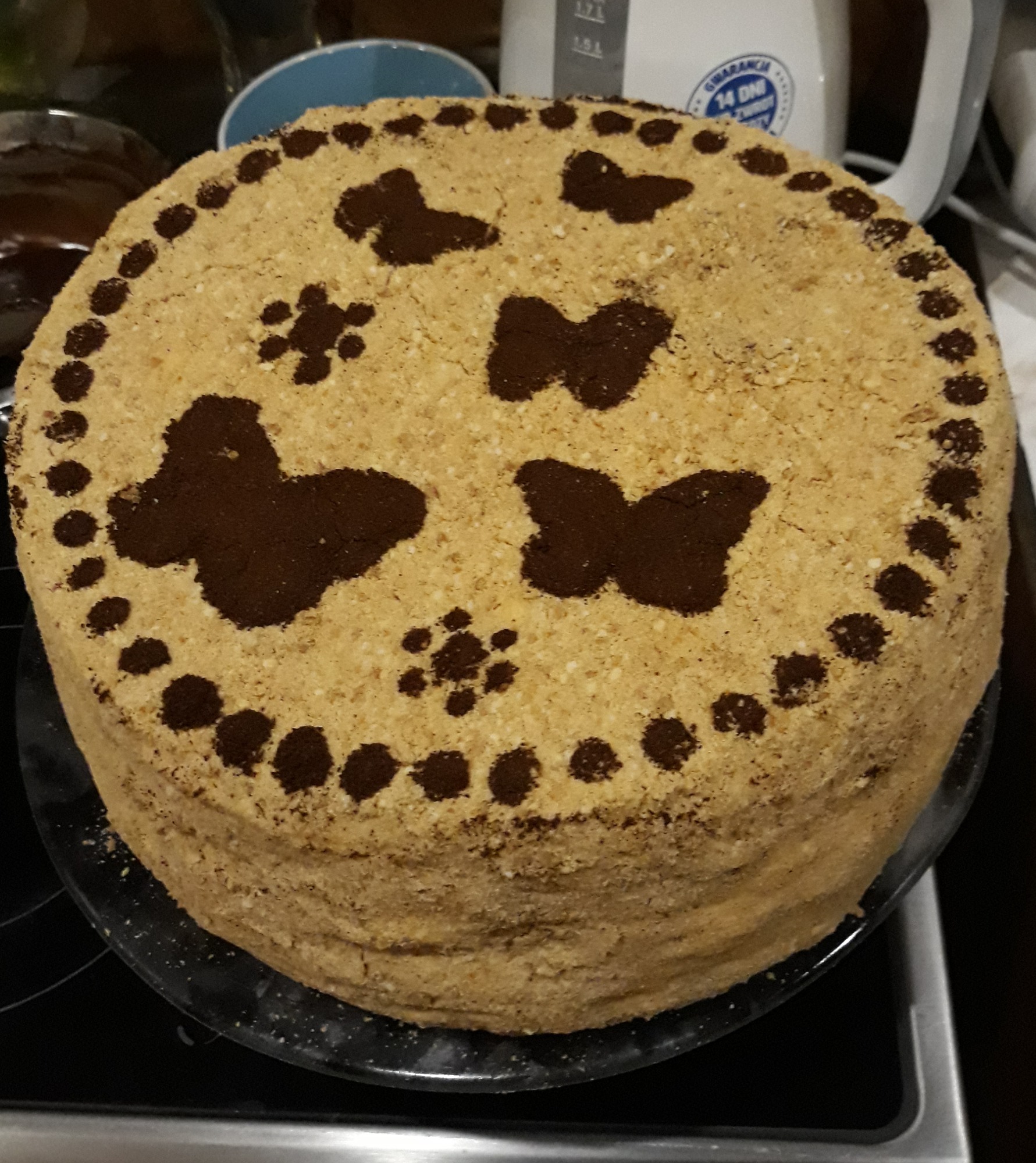 Wielowarstwowy tort z jasnym kremem, udekorowany motylami i kropkami z ciemnego kakao, umieszczony na szklanym talerzu.