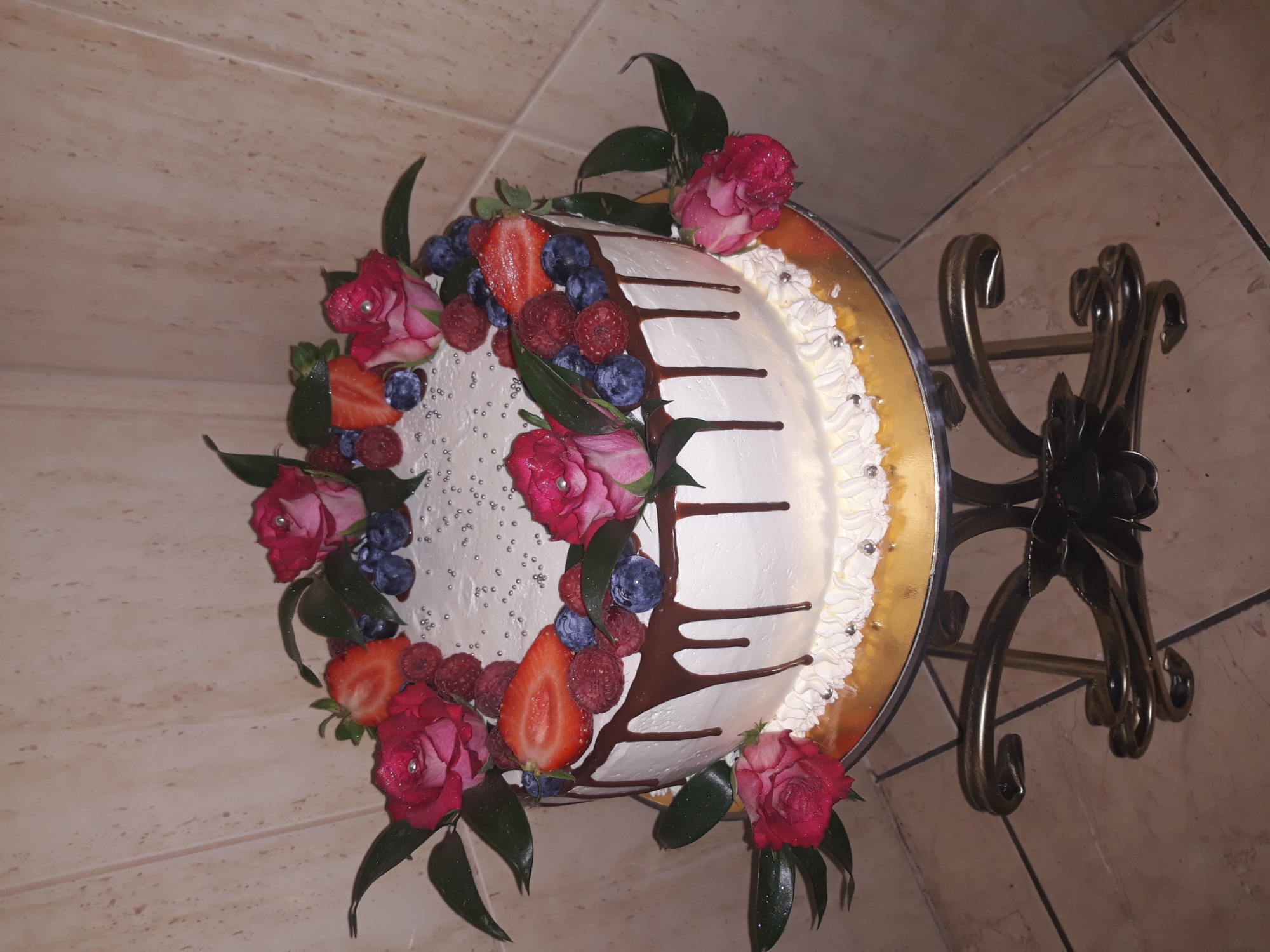 Tort z białym kremem, polany czekoladą i udekorowany świeżymi truskawkami, malinami, borówkami, różami oraz srebrnymi kuleczkami, umieszczony na metalowym stojaku.