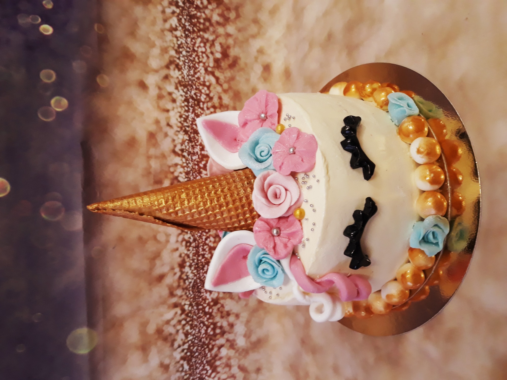 Tort jednorożec z kremową dekoracją z róż, złotym rogiem i lukrowanymi oczami na złotym podkładzie, ustawiony na tle z efektem bokeh.