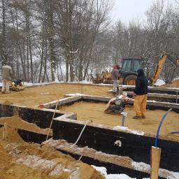 TROLEX - Budowa fundamentów w zimowej scenerii: robotnicy zagęszczają piasek w wykopie, w tle koparka i ośnieżone drzewa.