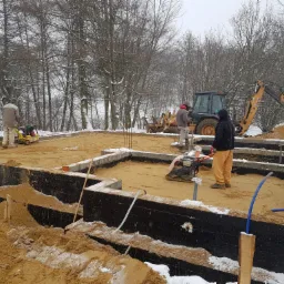 Budowa fundamentów w zimowej scenerii: robotnicy zagęszczają piasek w wykopie, w tle koparka i ośnieżone drzewa.