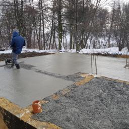 TROLEX - Wyrównywanie świeżo wylanego betonu na fundamencie domu jednorodzinnego przy użyciu zacieraczki mechanicznej, widoczne wystające pręty zbrojeniowe i zasypany śniegiem teren.