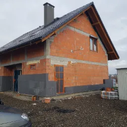 Dom jednorodzinny w trakcie budowy: ściany z czerwonej cegły częściowo ocieplone szarym styropianem, dach pokryty czarną dachówką, widoczne okna i komin, teren wokół domu w trakcie prac budowlanych.