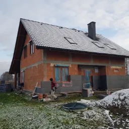 Budowa domu murowanego: robotnicy ocieplają elewację szarymi płytami styropianowymi w chłodny dzień z lekkim śniegiem na dachu i trawie.