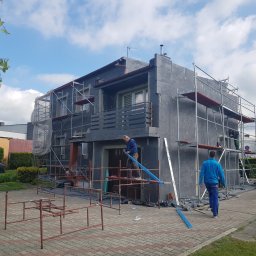 TROLEX - Dwukondygnacyjny dom jednorodzinny w trakcie ocieplania, obłożony szarymi płytami styropianowymi, otoczony rusztowaniami, na pierwszym planie pracownicy budowlani i rozłożone elementy rusztowania...