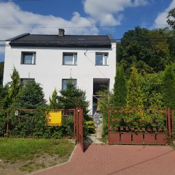 TROLEX - Dwukondygnacyjny dom z białymi ścianami i ciemnym dachem, otoczony zielenią, widoczny zza otwartej, brązowej bramy wjazdowej z ceglaną ścieżką.