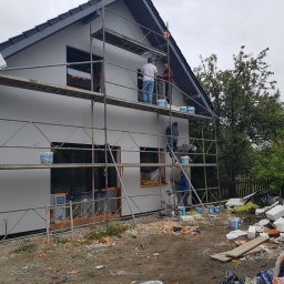 TROLEX - Pracownicy na rusztowaniu podczas nakładania tynku na elewację domu jednorodzinnego z szarą dachówką, widoczne wiadra z farbą i materiały izolacyjne na ziemi.