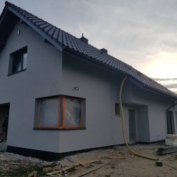 TROLEX - Nowo otynkowany dom jednorodzinny z szarą elewacją, czarnym dachem z dachówki ceramicznej, okna zabezpieczone folią malarską, widoczny wąż do tynkowania i bałagan na posesji w trakcie prac...