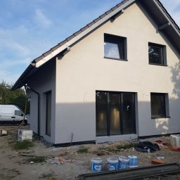 TROLEX - Nowo otynkowany dom jednorodzinny z szarą elewacją i ciemnymi oknami, z widocznym samochodem dostawczym firmy budowlanej w tle oraz materiałami budowlanymi na pierwszym planie.