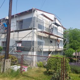 TROLEX - Prace elewacyjne w toku: kilku robotników na rusztowaniach, tynkowanie jasnej elewacji domu jednorodzinnego, okna zabezpieczone folią, widoczny fragment ogrodu z metalowym ogrodzeniem.