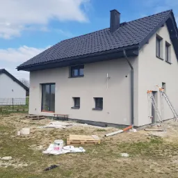 Nowoczesny dom jednorodzinny z szarą elewacją i ciemną dachówką, widoczne dwie drabiny opierające się o ścianę, teren wokół domu częściowo zagospodarowany, z paletami i resztkami materiałów...