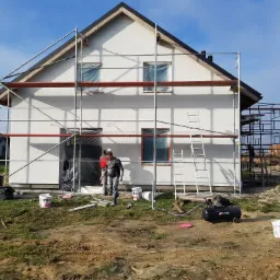 Nowo otynkowany dom jednorodzinny w trakcie prac wykończeniowych elewacji, z widocznymi rusztowaniami, zabezpieczonymi oknami folią, kompresorem i pracownikami w kombinezonach malarskich.