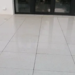 Błyszczący taras wyłożony jasnymi, kwadratowymi płytkami z delikatnym wzorem, przylegający do ściany budynku z czarnymi, przeszklonymi drzwiami balkonowymi.