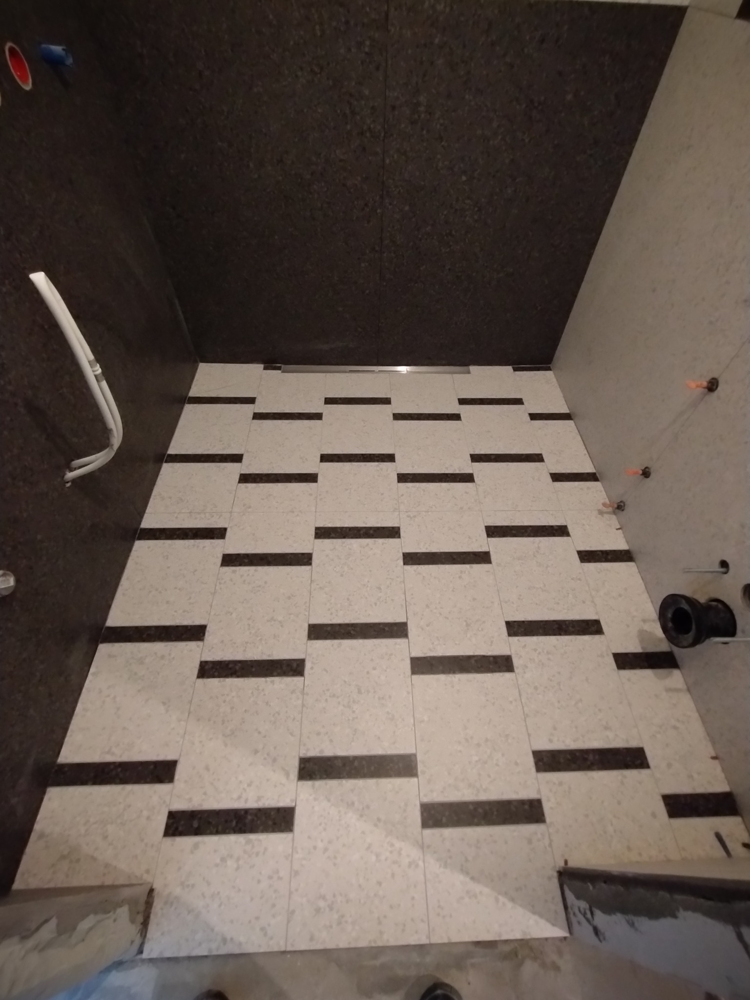 Fragment łazienki w trakcie prac wykończeniowych: ułożona podłoga z jasnych płytek z ciemnymi paskami, ciemna okładzina ścienna, widoczne elementy instalacji hydraulicznej i poziomowania płytek.