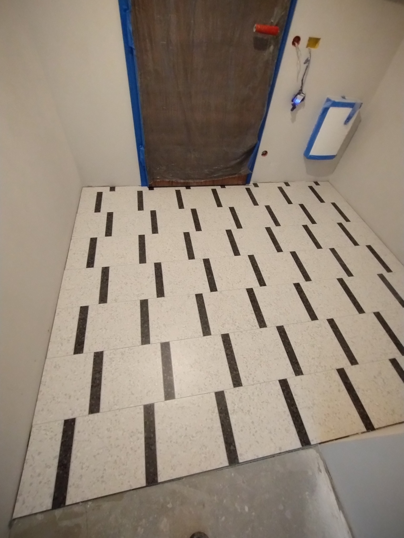 Ułożone płytki podłogowe w łazience z wzorem białych płytek z czarnymi paskami, zabezpieczone folią drzwi i fragment instalacji elektrycznej na ścianie.