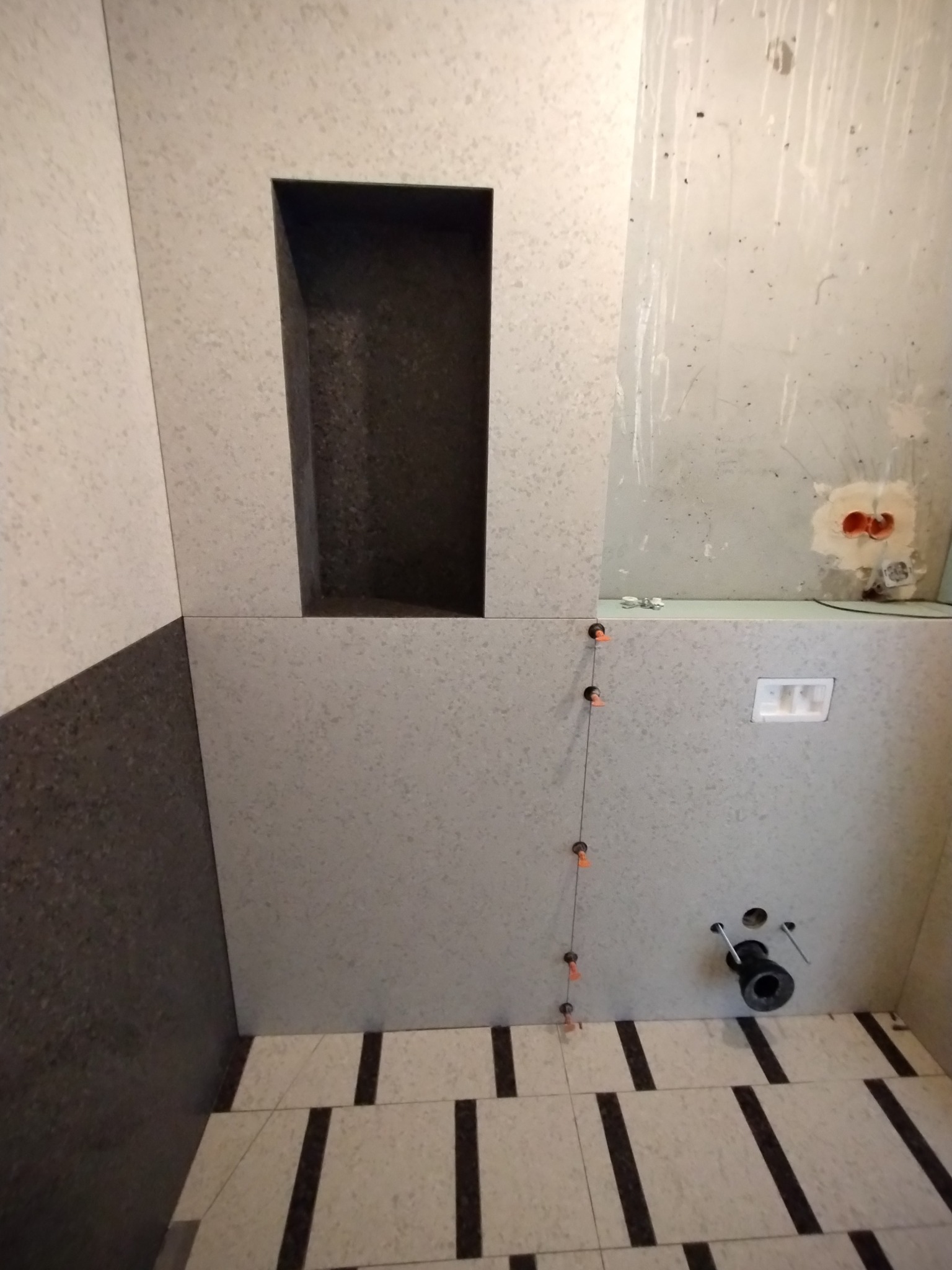 Ściana łazienki w trakcie układania płytek z wnęką, widoczne elementy instalacji wodno-kanalizacyjnej i krzyżyki dystansowe.