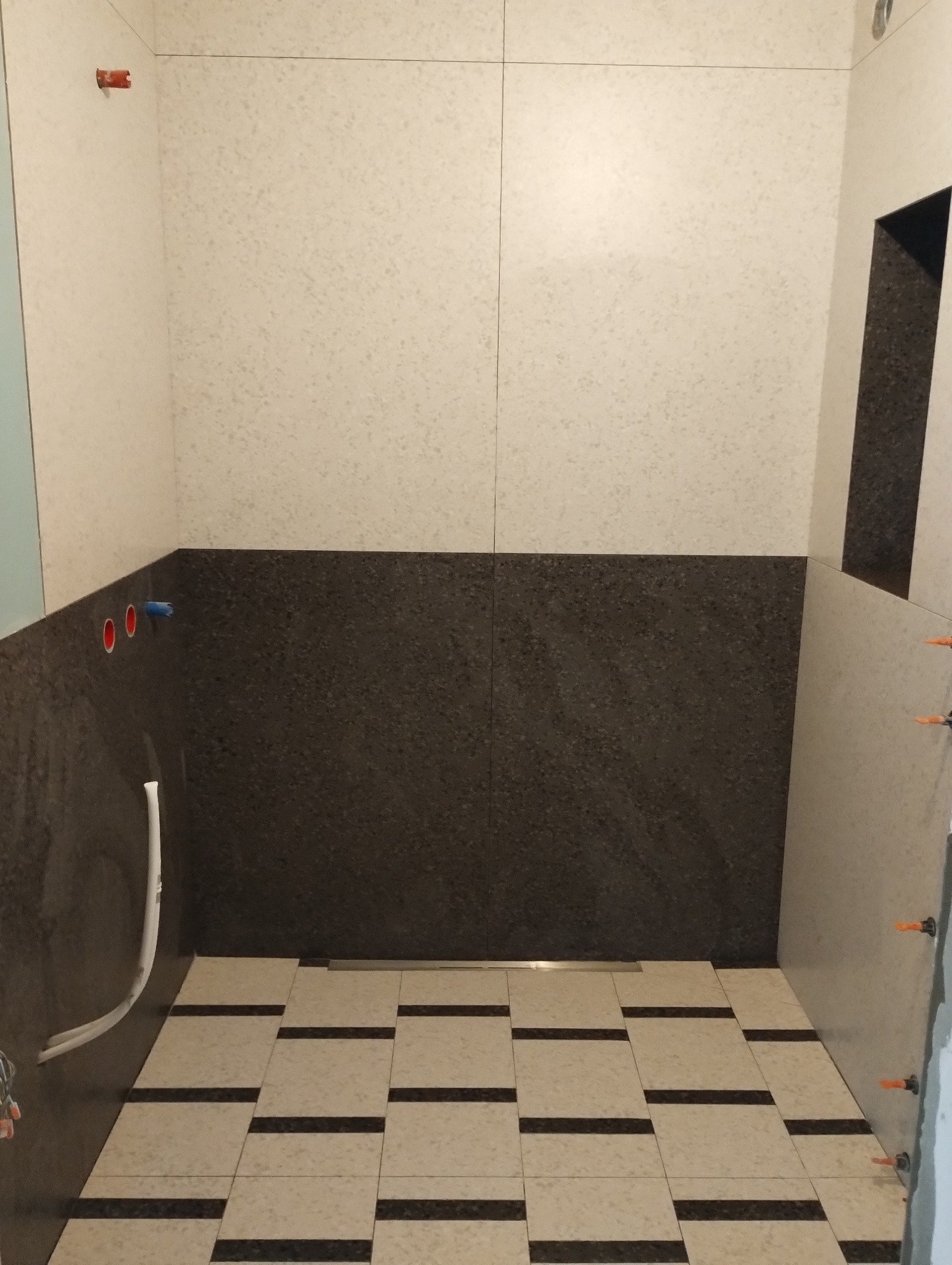 Wnętrze łazienki w trakcie remontu: dolna część ścian wyłożona ciemnymi płytkami, górna jasnymi, na podłodze płytki w pasy, widoczne rury i poziomica na ścianie.