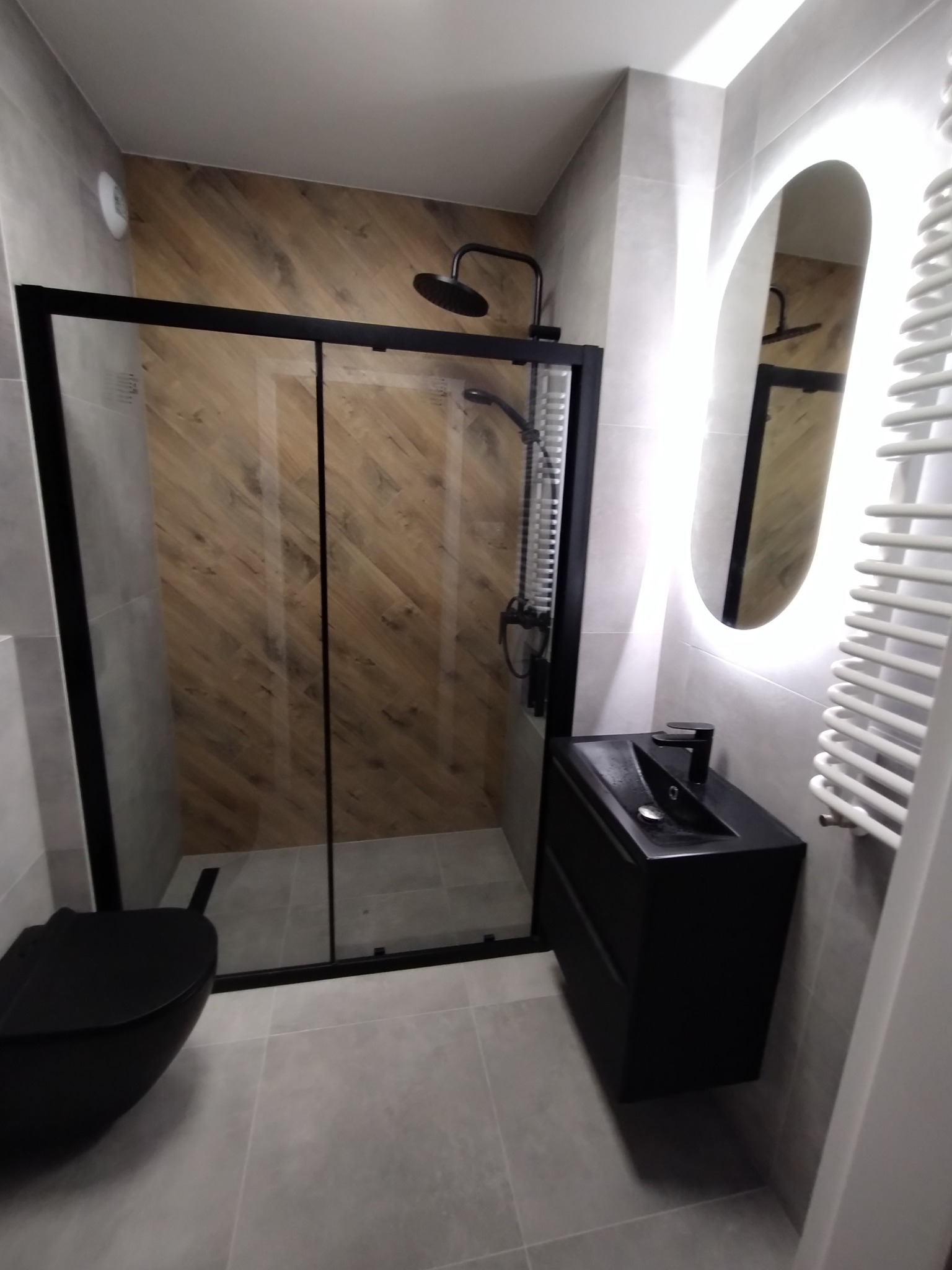 Nowoczesna łazienka z czarną armaturą: kabina prysznicowa z drewnopodobnymi płytkami, umywalka z szafką i podświetlane lustro owalne.