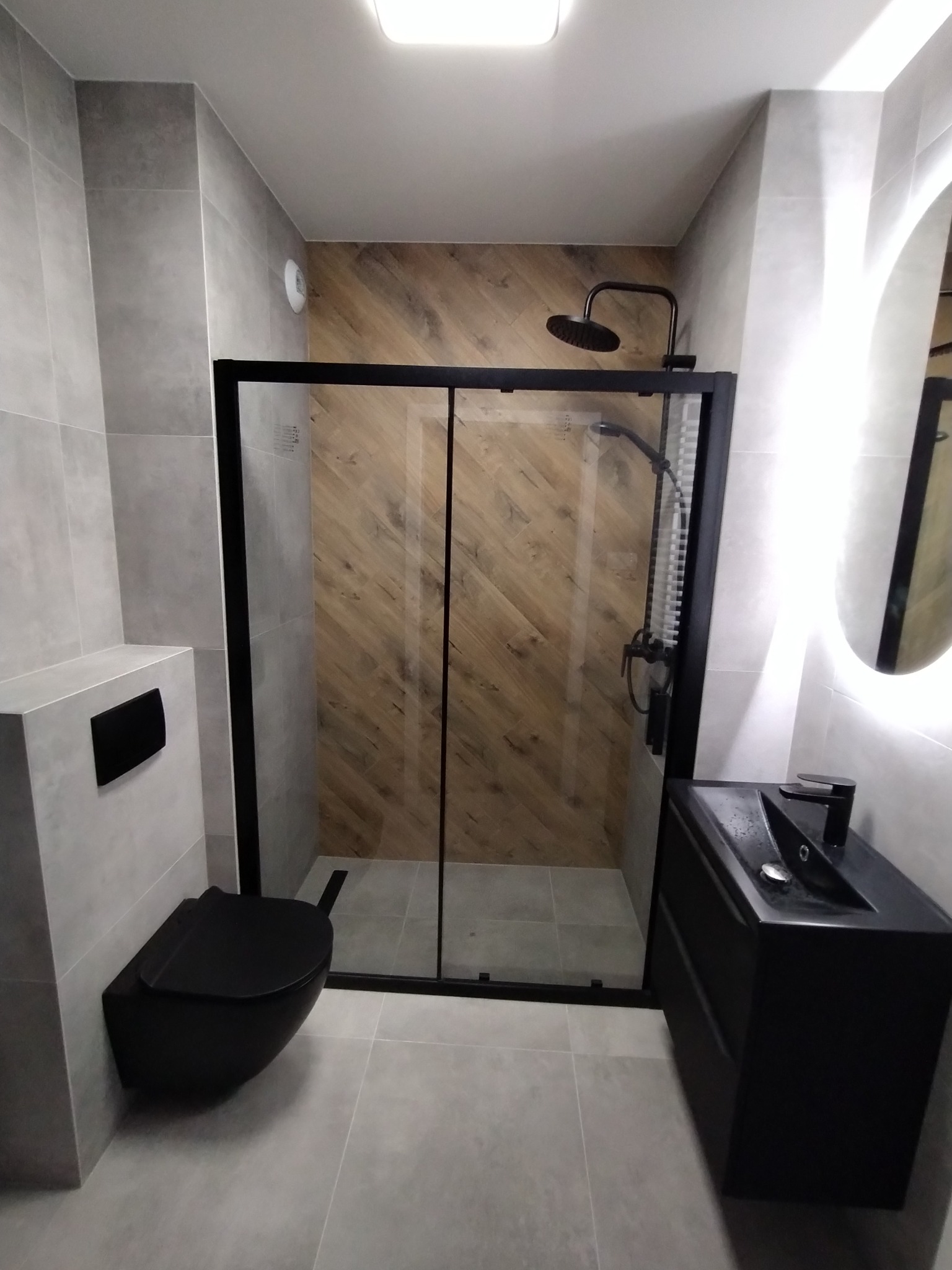 Nowoczesna łazienka z szarymi płytkami, czarną armaturą, kabiną prysznicową z drewnopodobną ścianą i podświetlanym lustrem.