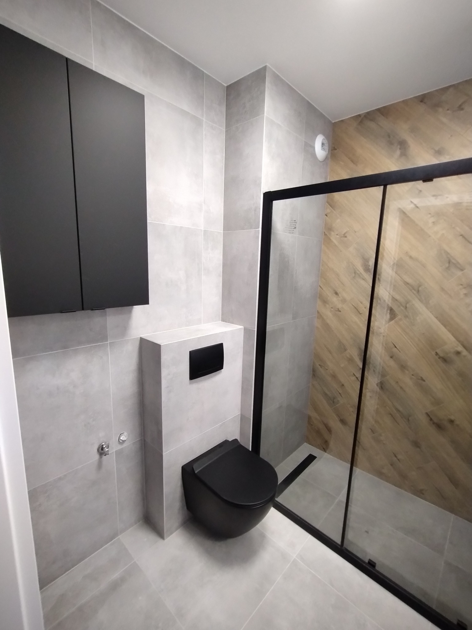 Nowoczesna łazienka z szarymi płytkami, czarną armaturą i kabiną prysznicową z drewnopodobnymi panelami na ścianie.