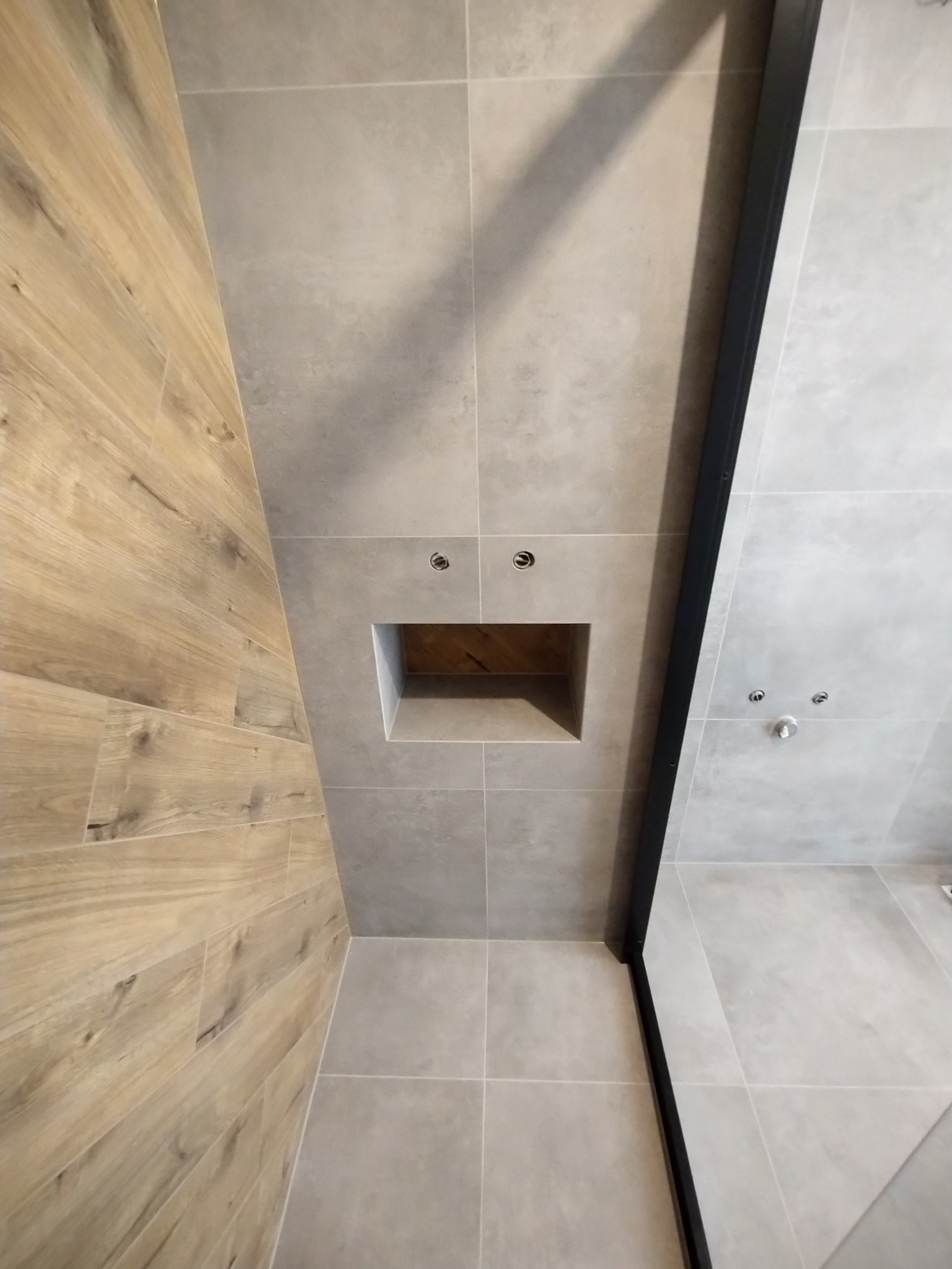 Nowoczesna łazienka z drewnopodobnymi płytkami na ścianie, szare płytki ceramiczne, wnęka w ścianie wyłożona drewnem i czarna rama kabiny prysznicowej.