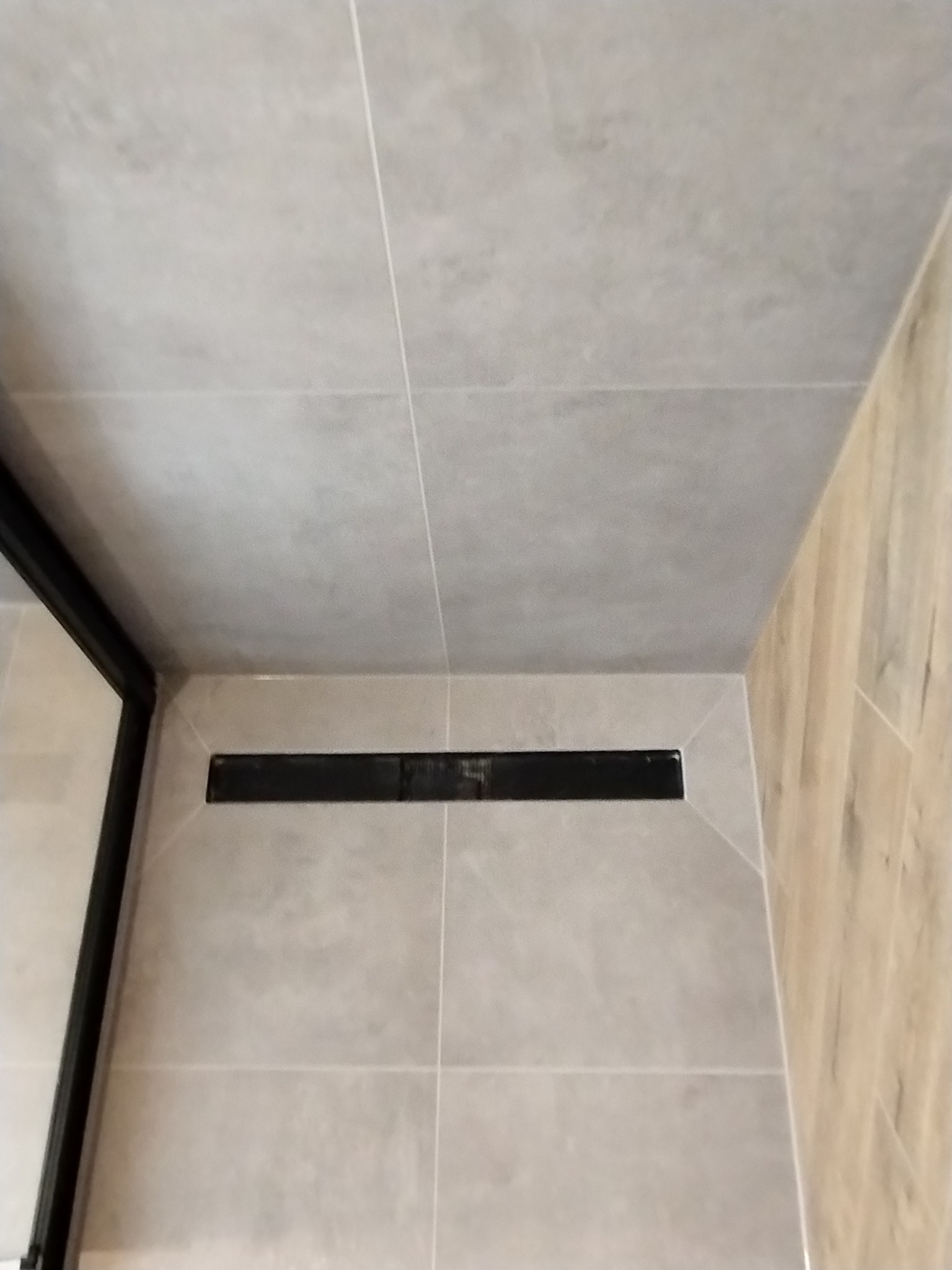 Szczegół wykończenia łazienki: odpływ liniowy w posadzce wyłożonej płytkami ceramicznymi w odcieniach szarości, z fragmentem kabiny prysznicowej z czarną ramą.