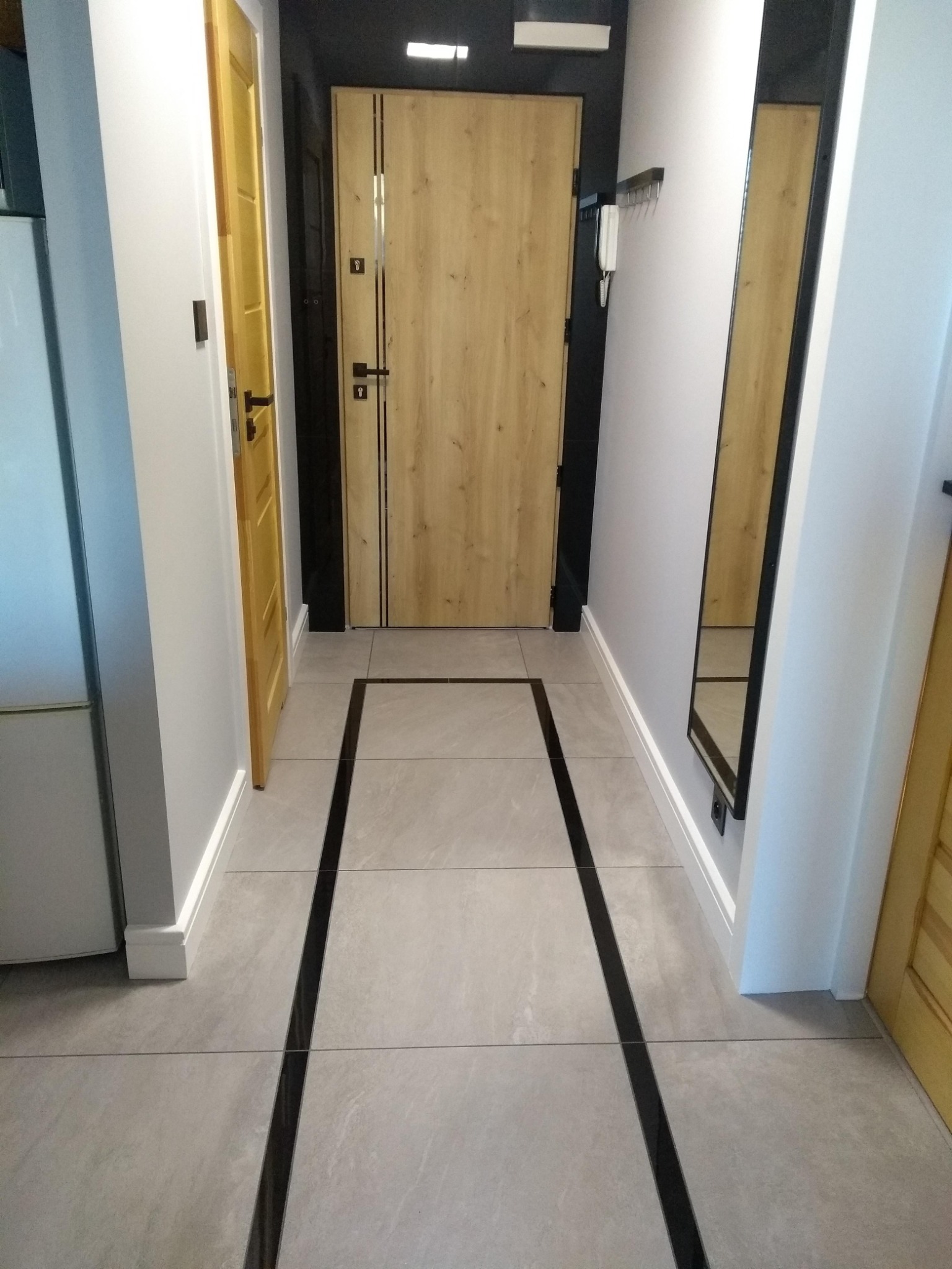 Wykończony korytarz z jasnymi płytkami podłogowymi z czarnymi akcentami, dwoje drzwi w kolorze drewna, lustro w czarnej ramie i szare ściany.