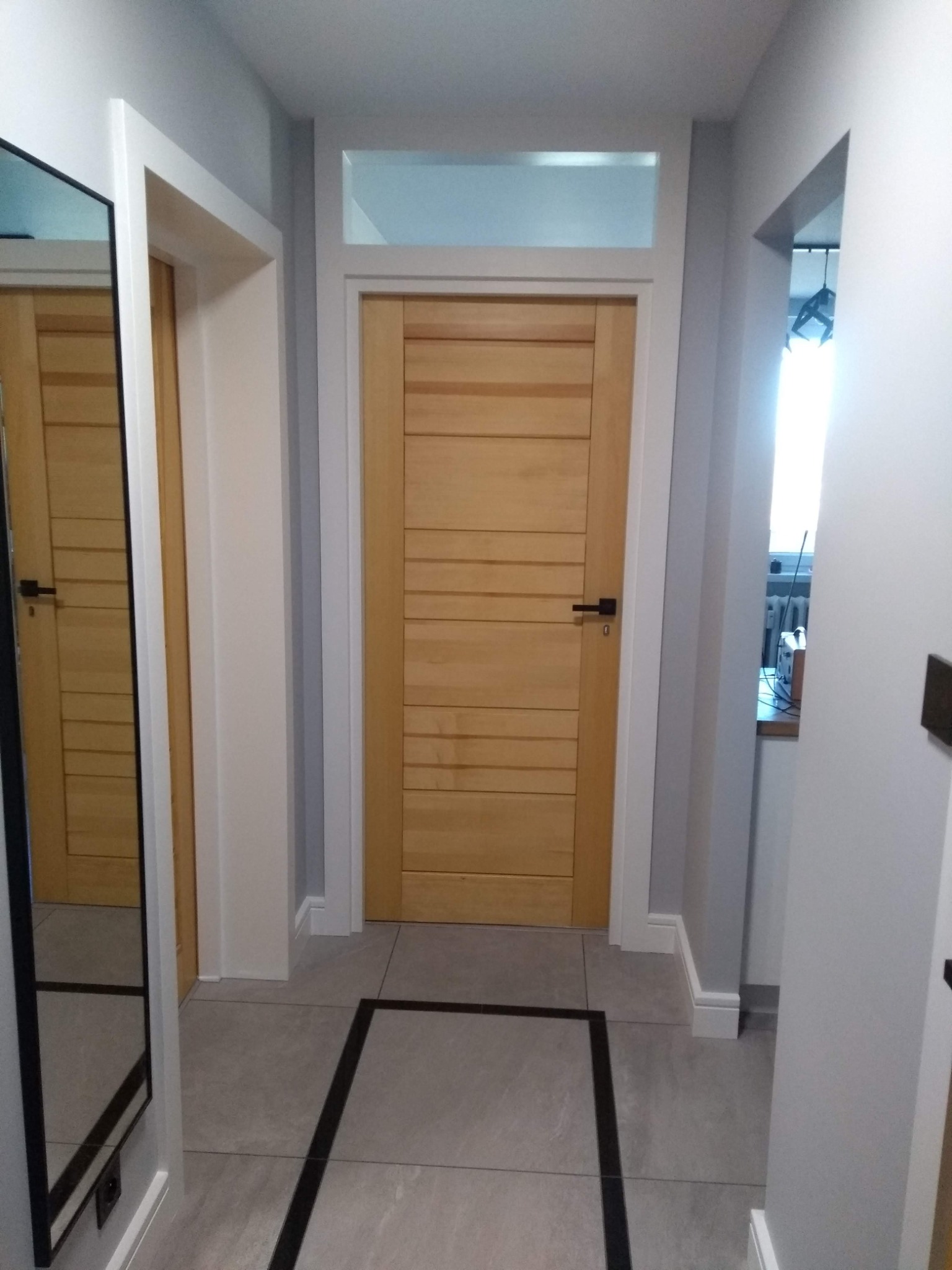 Korytarz z dwiema drewnianymi drzwiami z poziomymi panelami, szarą podłogą z ciemnymi akcentami, białe ościeżnice i szare ściany, widoczne fragmenty lustra i lampy w tle.