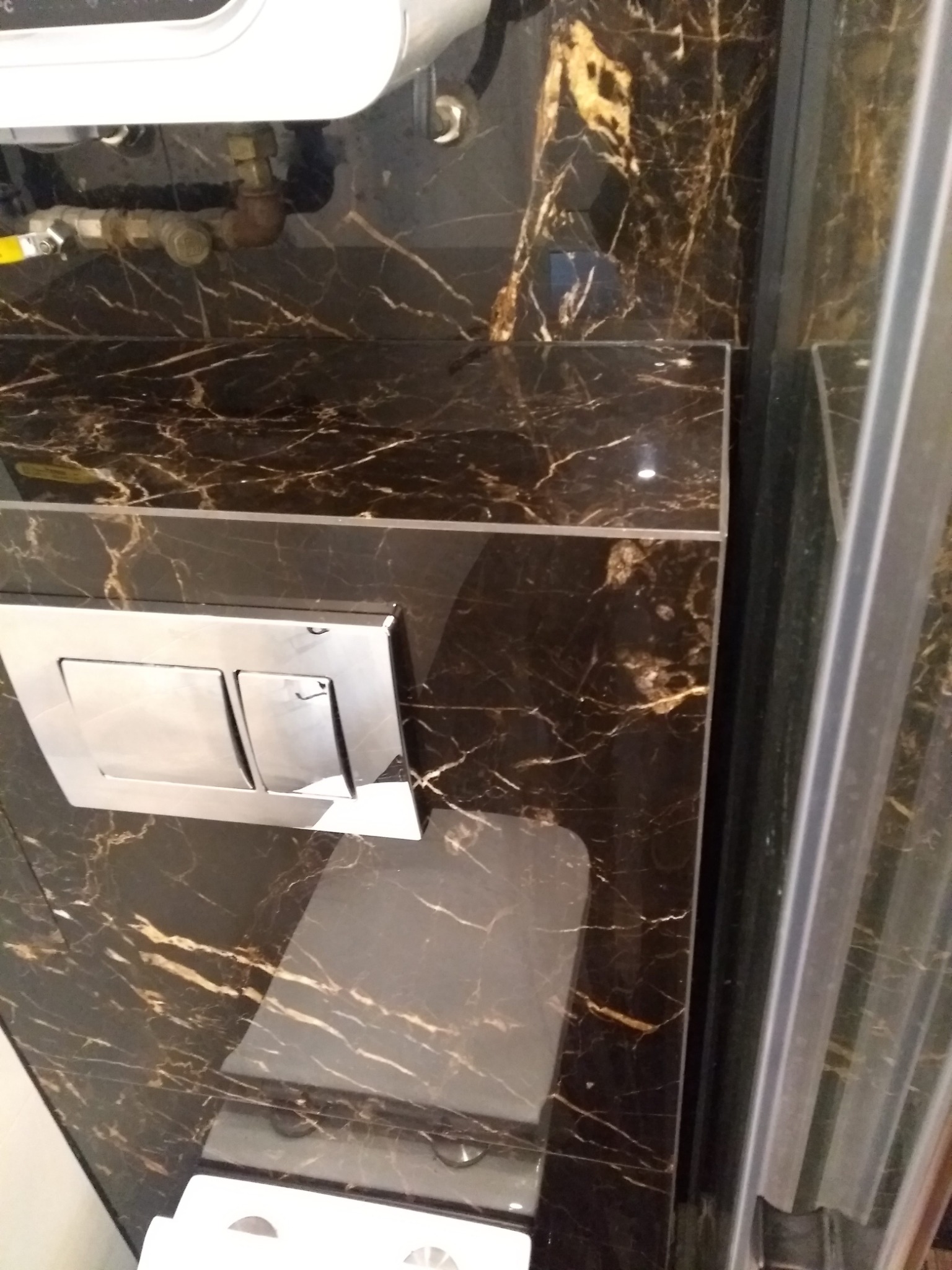 Czarny marmur z złotymi żyłami na ścianie łazienki, widoczny chromowany przycisk spłuczki i fragment kabiny prysznicowej.