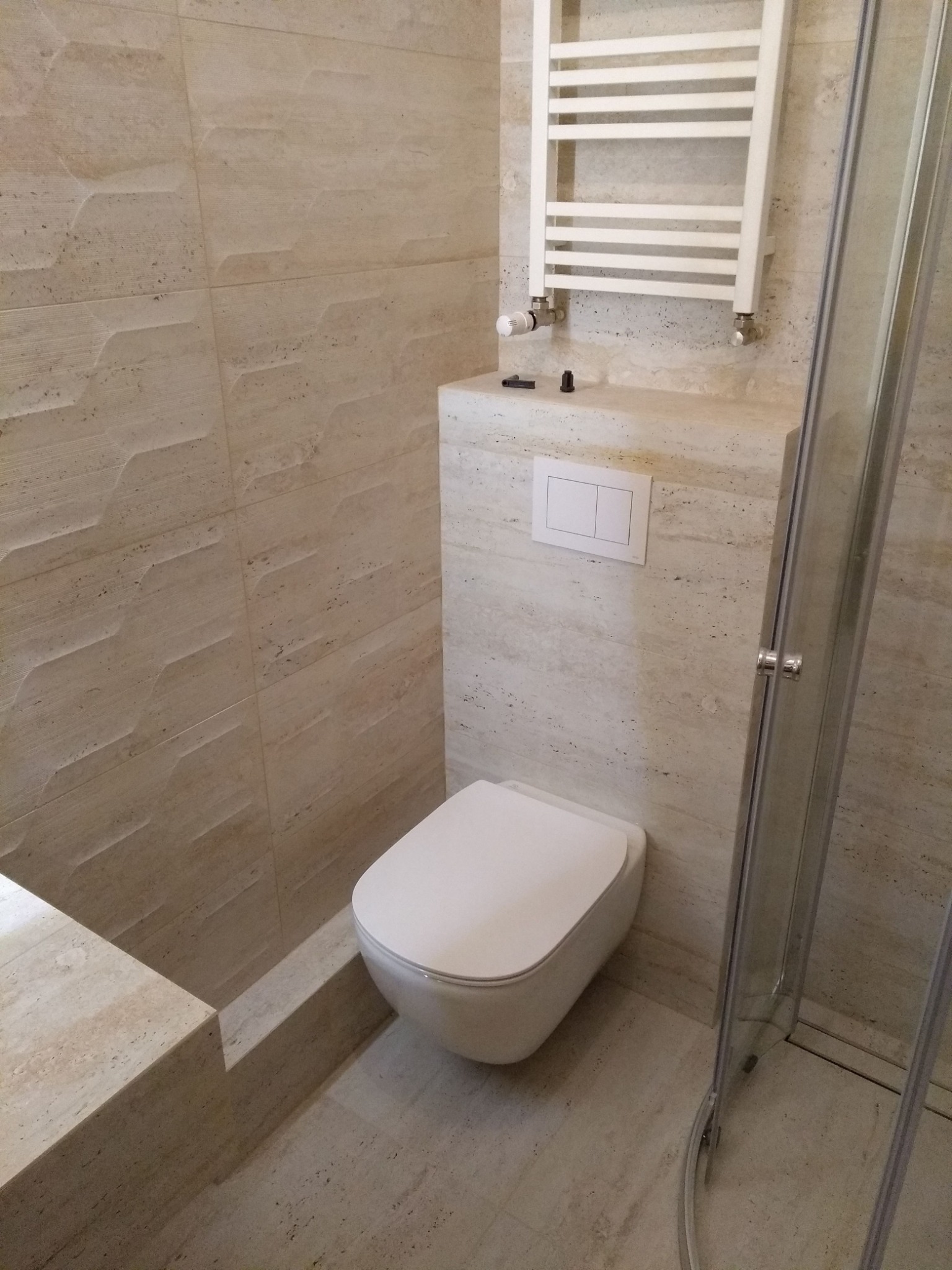 Beżowa łazienka z toaletą podwieszaną, grzejnikiem drabinkowym i kabiną prysznicową ze szklanymi drzwiami, płytki z delikatnym wzorem.