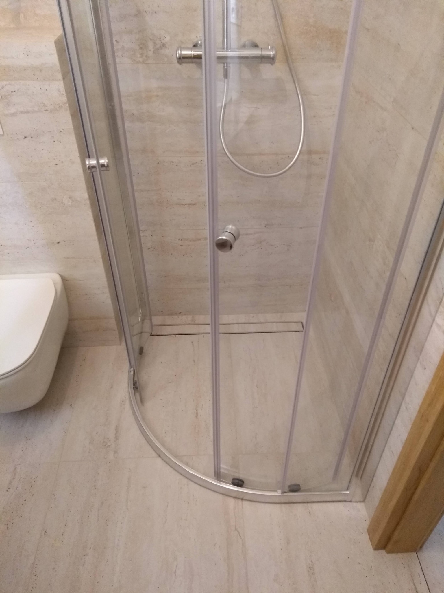 Nowoczesna kabina prysznicowa z zaokrąglonymi drzwiami, zamontowana w narożniku łazienki wyłożonej beżowymi płytkami imitującymi kamień, z widocznym odpływem liniowym i wiszącą miską WC po lewej...