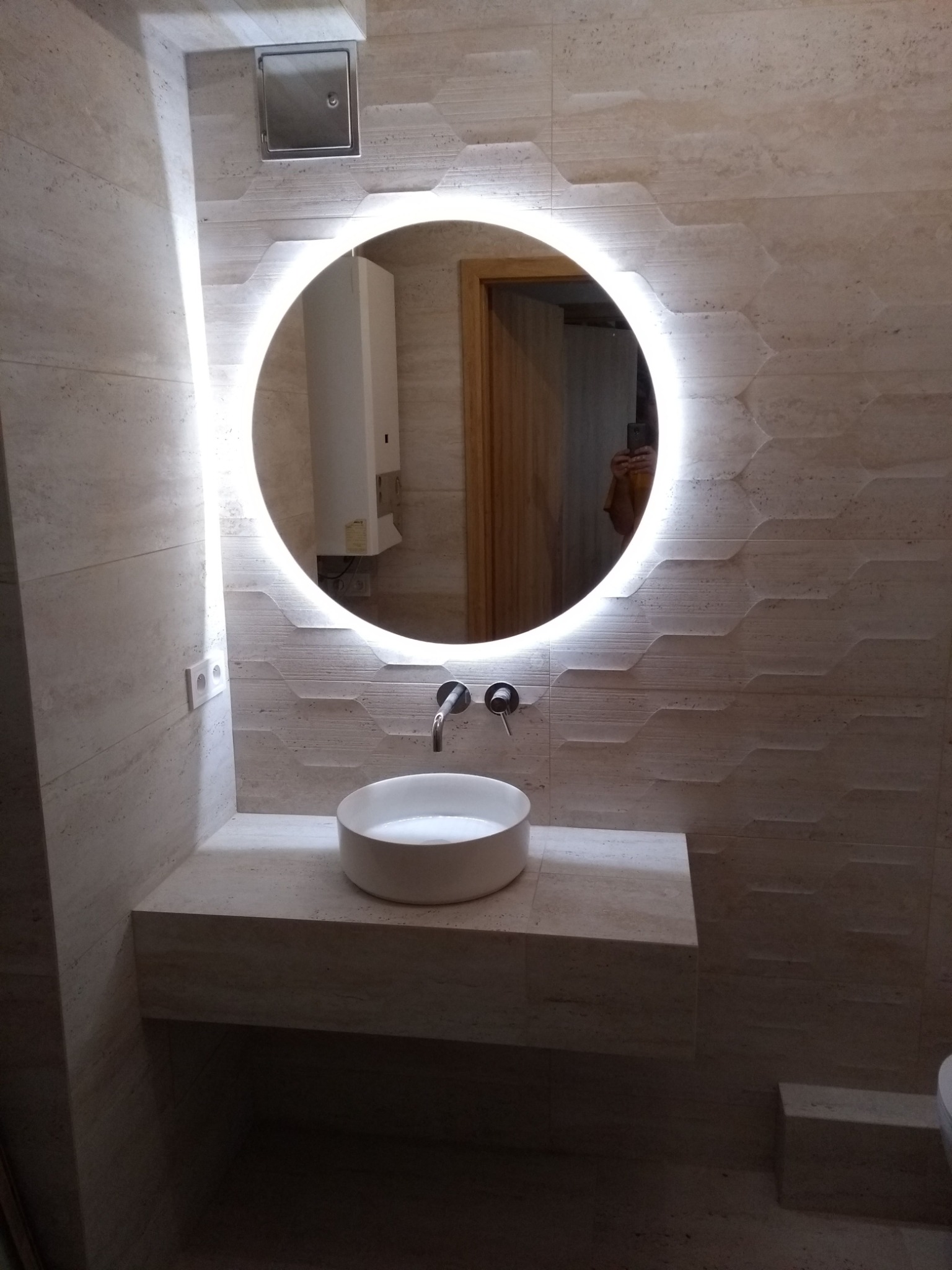 Nowoczesna łazienka z okrągłym lustrem podświetlanym LED, umywalką nablatową i płytkami imitującymi trawertyn o geometrycznym wzorze.