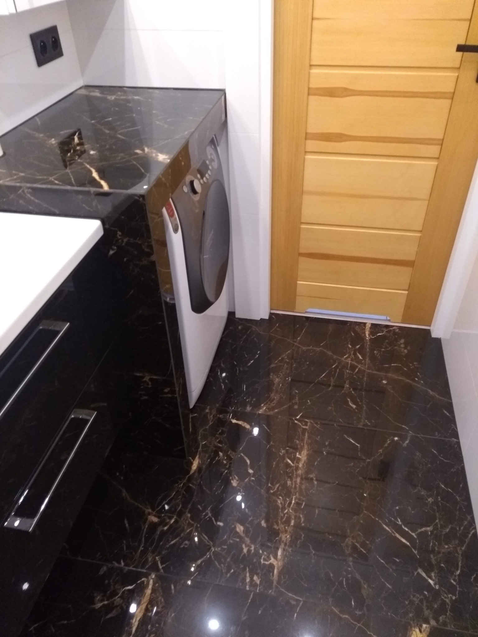 Ciemna, marmurkowa podłoga i blat w łazience z drewnianymi drzwiami i białą pralką.