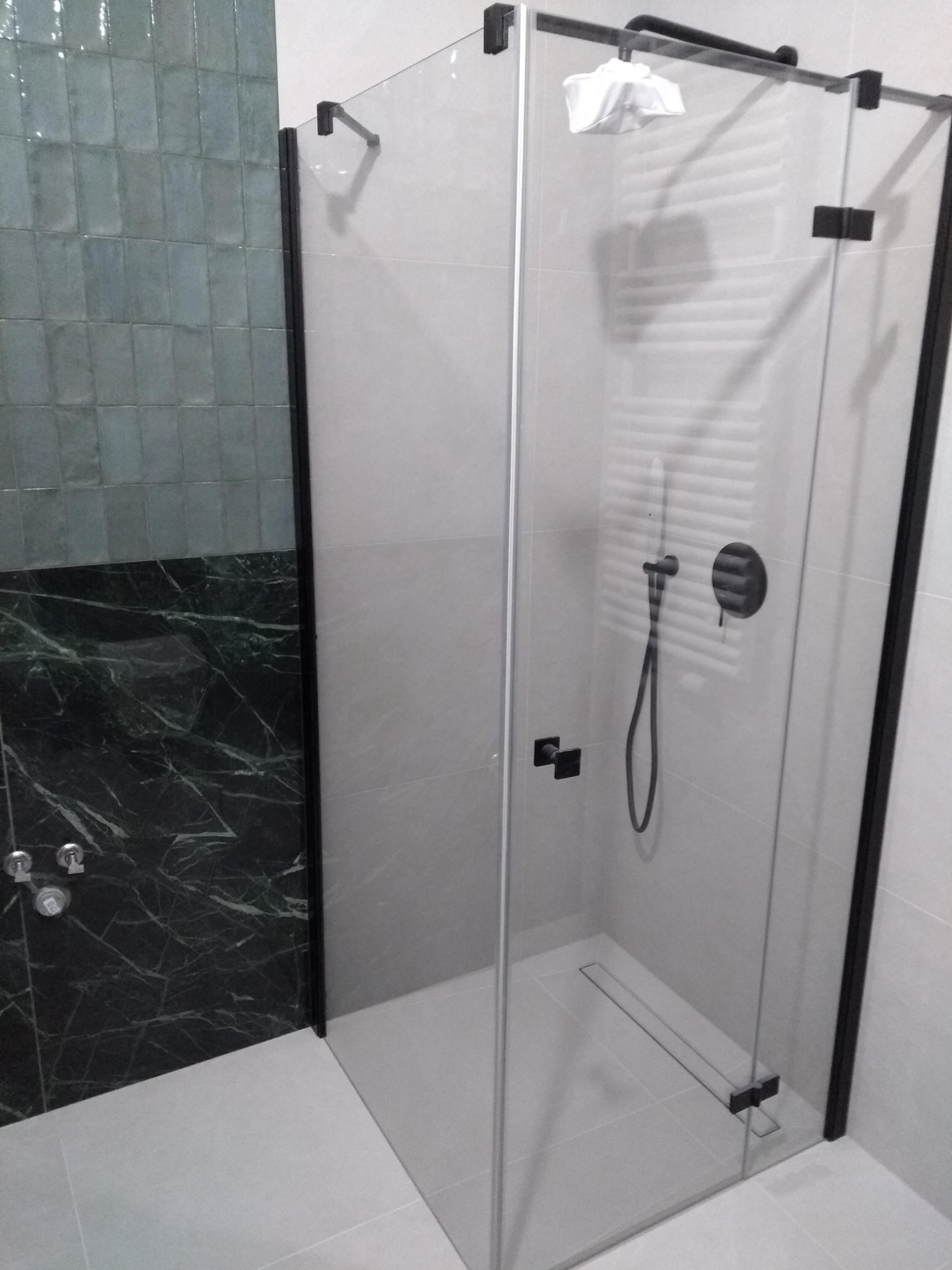 Kabina prysznicowa z czarnymi profilami, widoczny odpływ liniowy, bateria podtynkowa i zielona ściana z płytek.