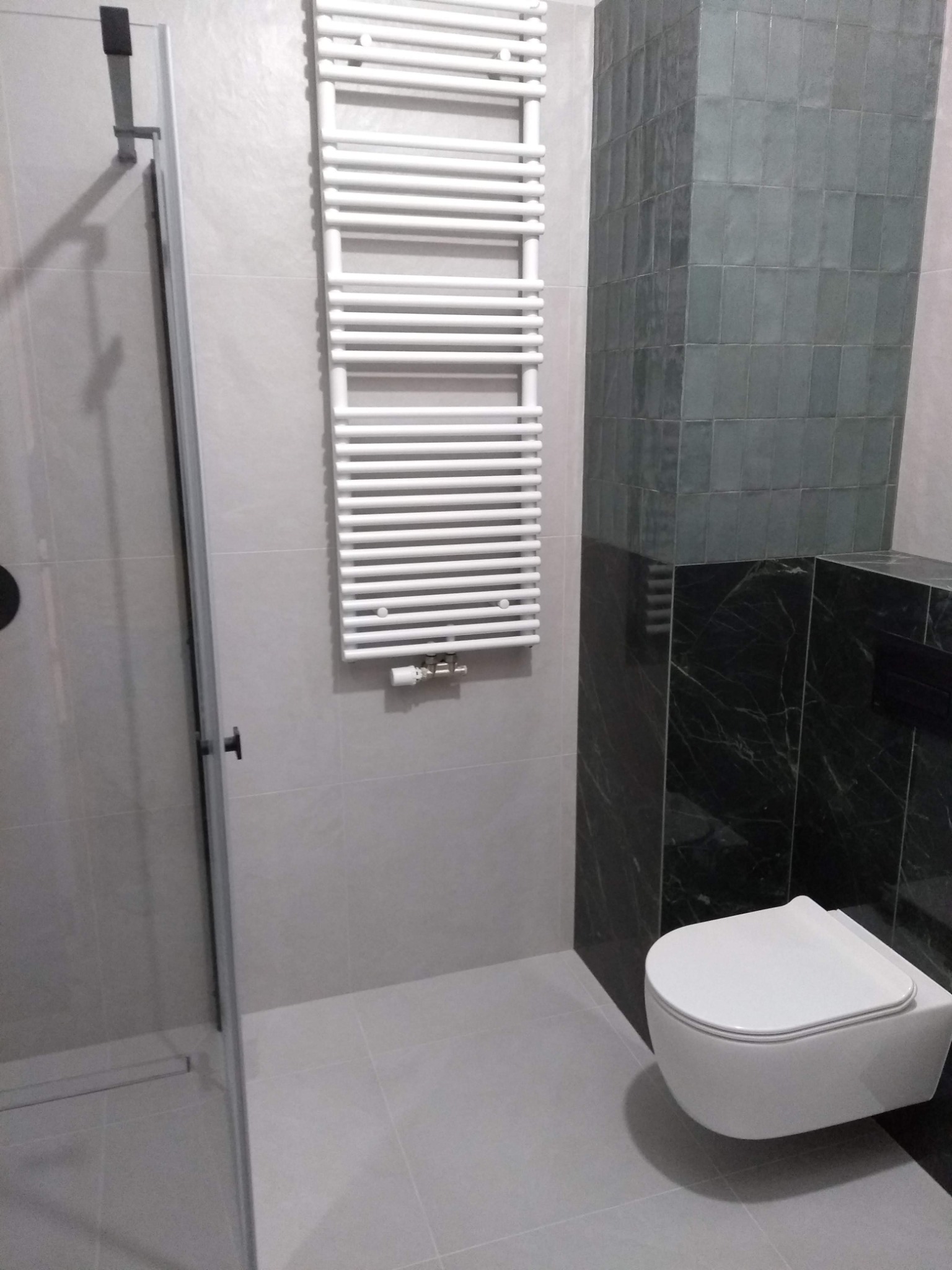 Nowoczesna łazienka z kabiną prysznicową, grzejnikiem drabinkowym i podwieszaną toaletą, wykończona szarymi i zielonymi płytkami.