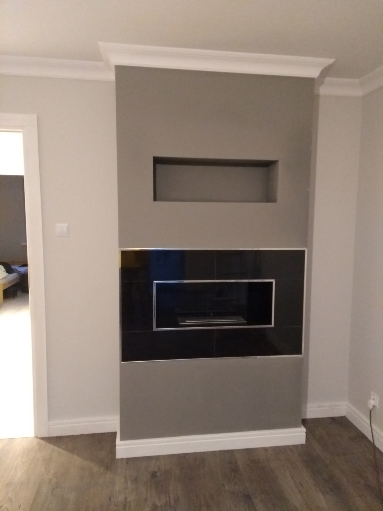 Szara ściana z wnęką i czarną, błyszczącą obudową kominka z płytek, wykończona białymi listwami przypodłogowymi i sufitowymi.