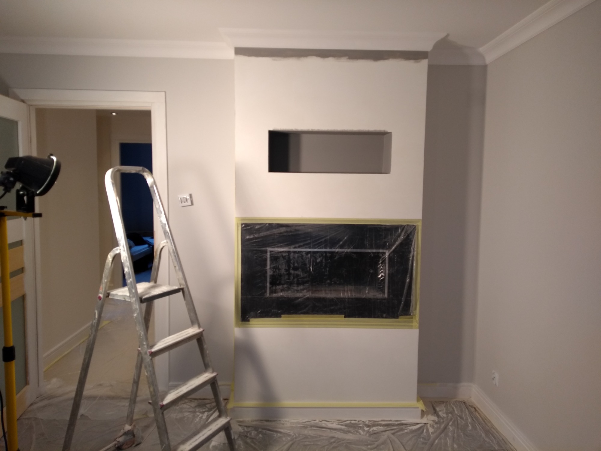 Ściana w trakcie przygotowania do malowania, z zabezpieczonym otworem na kominek folią i taśmą malarską, widoczna drabina i lampa oświetleniowa.