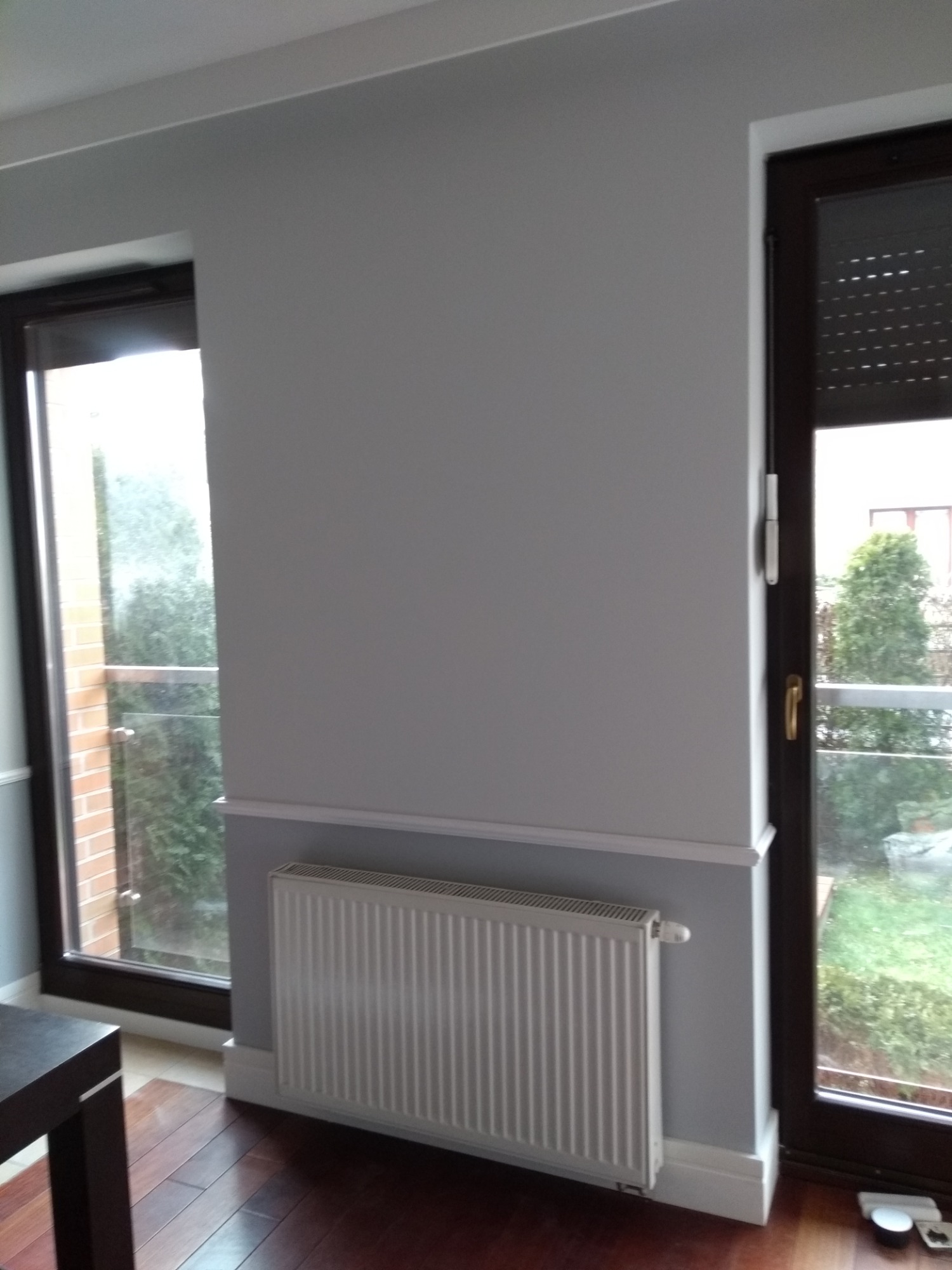 Szara ściana z grzejnikiem i dwoma oknami z ciemnymi ramami, widok na zewnątrz z zielenią, drewniana podłoga.