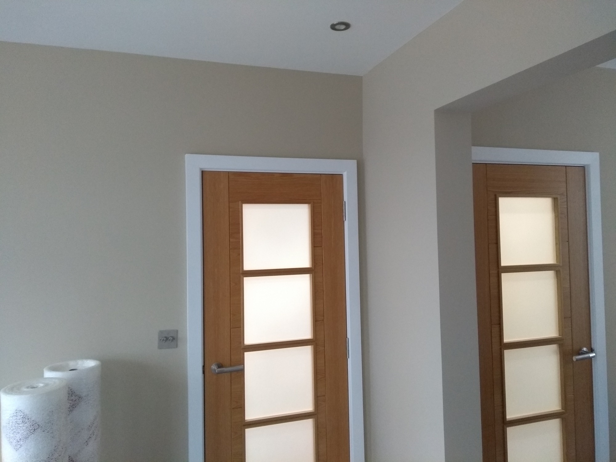 Dwa drewniane drzwi wewnętrzne z białymi ramami i przeszkleniami, widoczne rolki folii ochronnej na podłodze, ściany w odcieniach beżu.