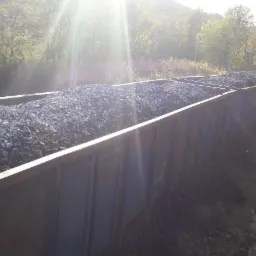 Wagony kolejowe wypełnione jasnoszarym węglem kamiennym w promieniach słońca, tło zalesione wzgórze.