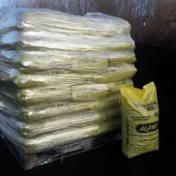 Stos worków z ekogroszkiem na palecie, owinięty folią stretch, obok pojedynczy worek z nadrukiem nazwy produktu i logo firmy, na tle czarnej pryzmy węgla i zabudowań.