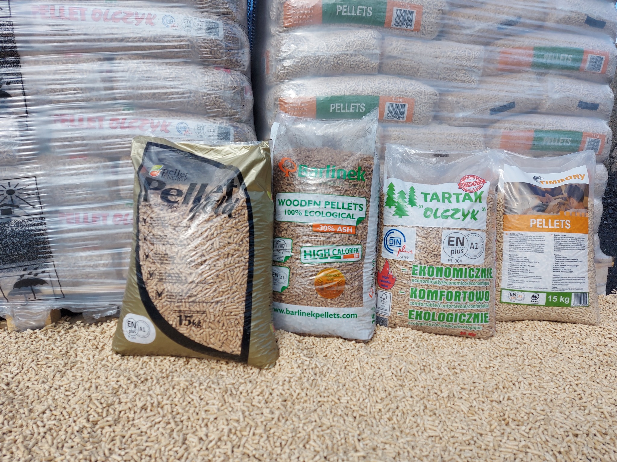 Ułożone worki z pelletem różnych producentów, w tym Barlinek, Tartak Olczyk i Timbory, na tle palet z zapasem pelletu w przezroczystej folii.