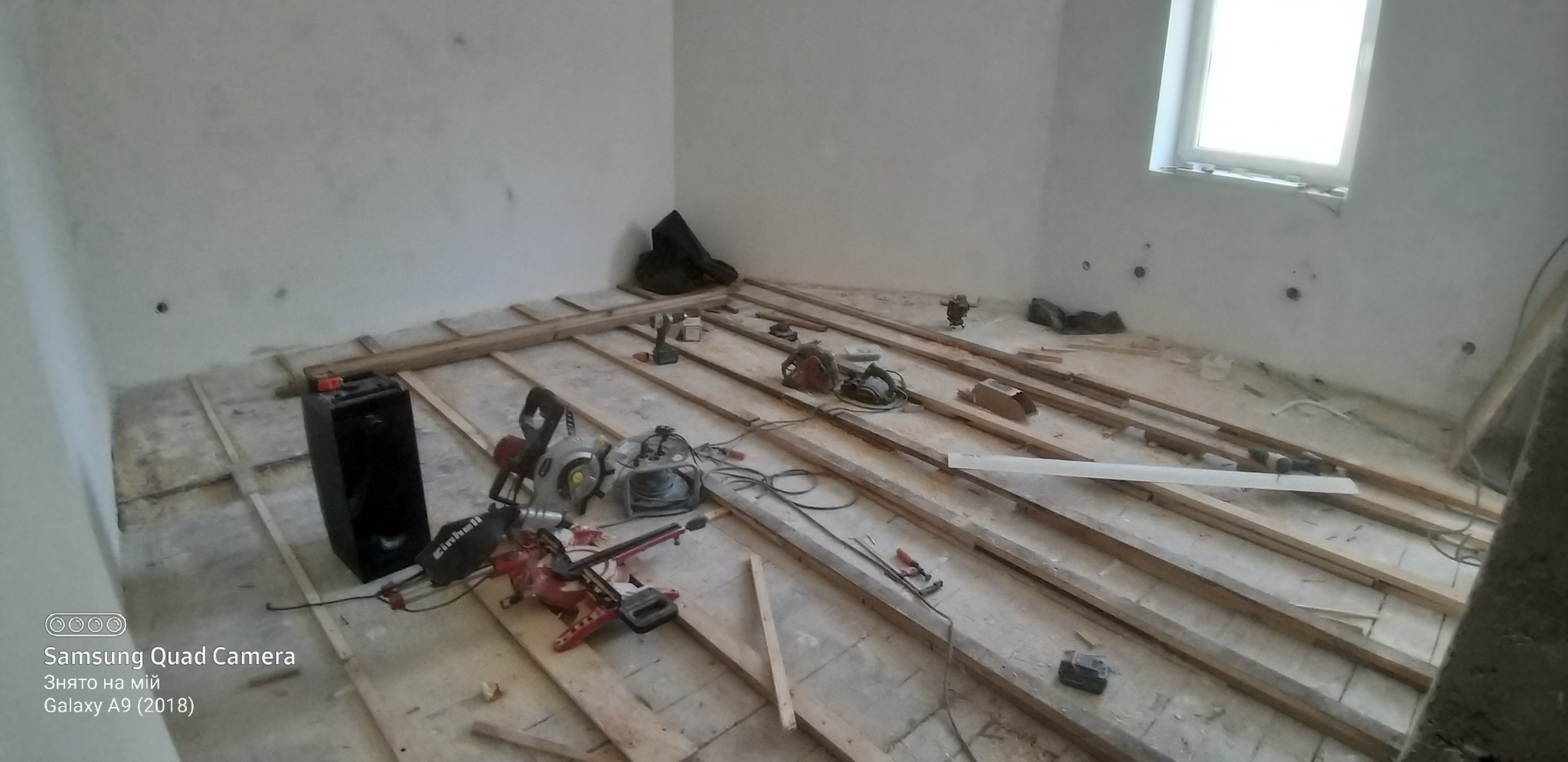 Przygotowanie podłoża pod podłogę w pokoju z oknem, widoczne drewniane legary, narzędzia: piła ukośnica, wiertarka, szlifierka, oraz głośnik przenośny.