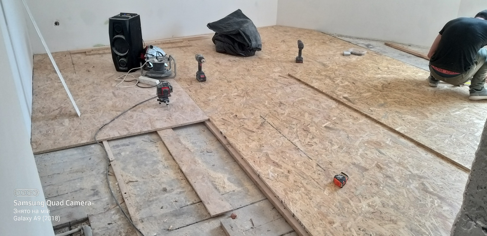 Układanie płyt OSB na podłodze w trakcie remontu, widoczny laser krzyżowy, wkrętarki, głośnik i częściowo ułożone płyty na nierównej podłodze.