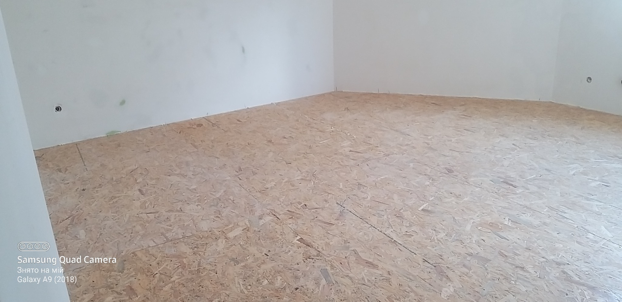 Surowe wnętrze pokoju z podłogą wyłożoną płytami OSB, ściany w trakcie przygotowania do malowania, widoczne puszki elektryczne.