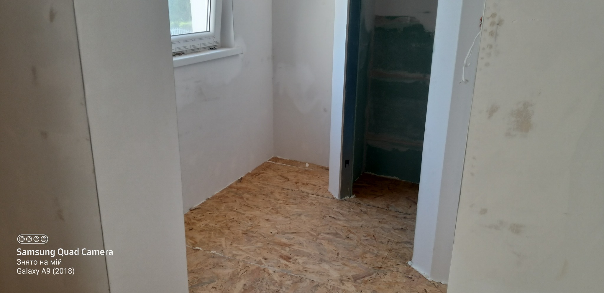 Surowe wnętrze w trakcie prac wykończeniowych: ściany po wstępnym szpachlowaniu, podłoga wyłożona płytami OSB, widoczne przygotowane wejście do garderoby z surowymi ścianami.