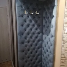 Wyściełana, pikowana alcowa garderoba w odcieniach szarości z trzema metalowymi haczykami, wkomponowana w przedpokój z przesuwną szafą.