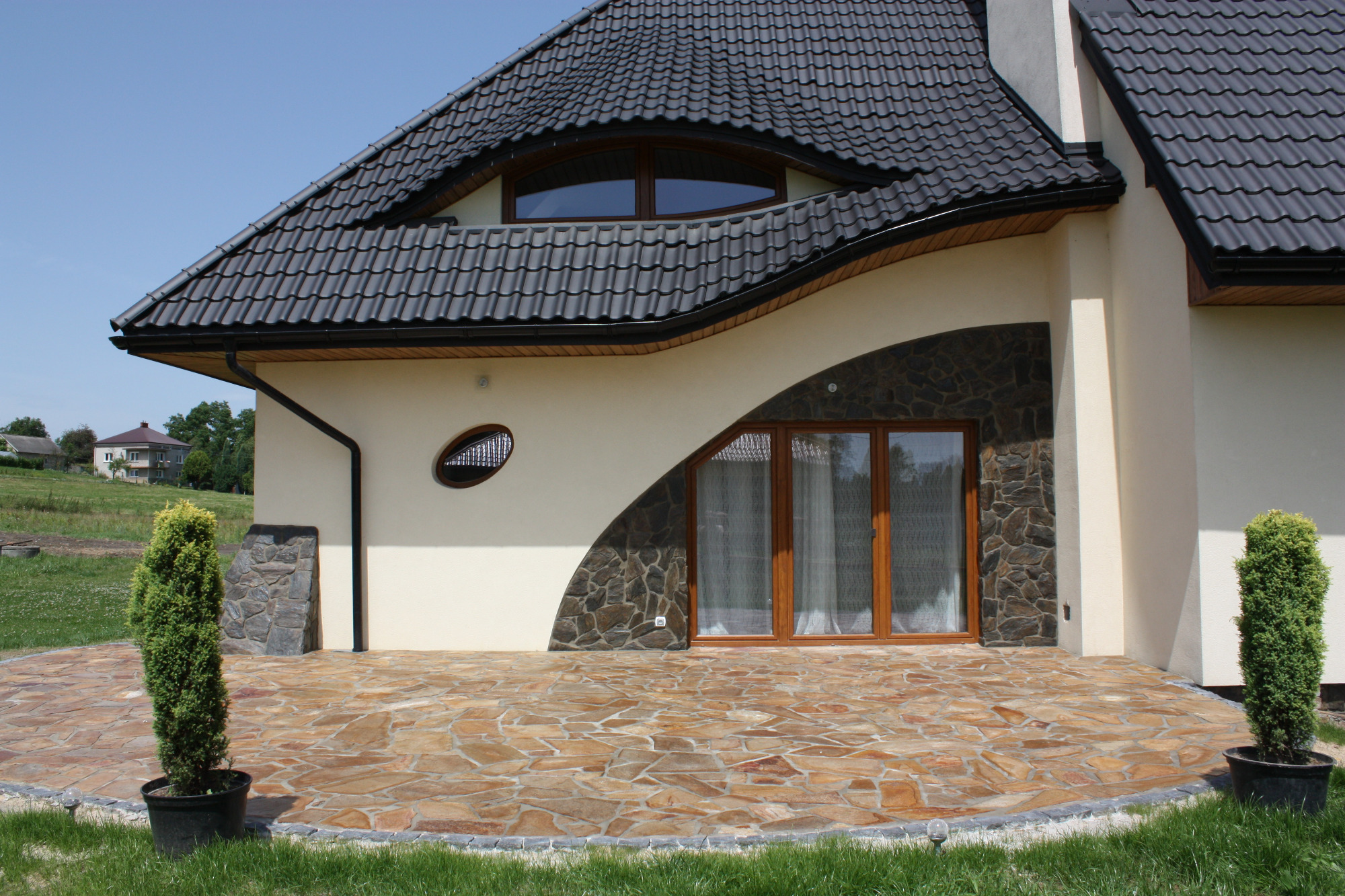 Taras z nieregularnych płyt kamiennych w odcieniach brązu i beżu, otoczony zielonym trawnikiem, przylegający do ściany domu z oknami o nietypowym kształcie i kamienną okładziną.